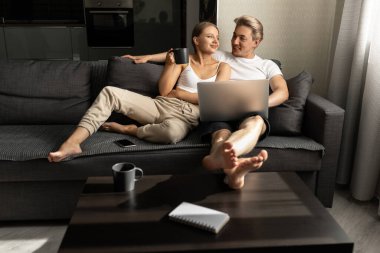 Güzel bir kadın ve bir erkek kanepede oturmuş sohbet ediyor, çay içiyor ve dizüstü bilgisayarda film izliyorlar.