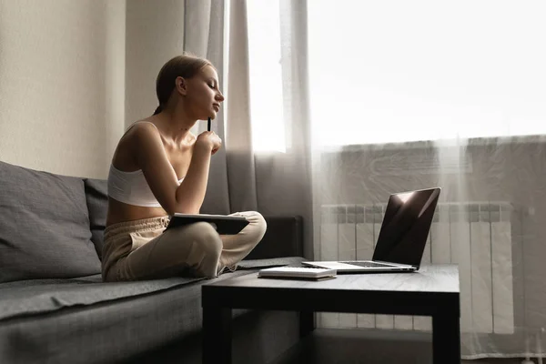 Belle femme caucasienne en vêtements décontractés assis sur le canapé dans l'appartement et ne fonctionne sur l'ordinateur, réfléchi — Photo