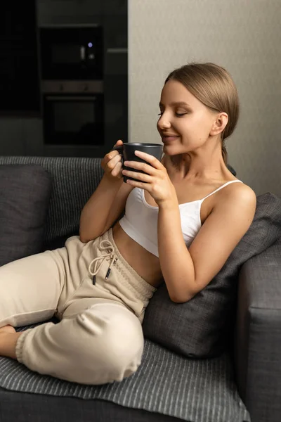 Krásná žena v horní části a kalhoty sedí na pohovce s hrnkem čaje a čichá k jeho lahodné aroma. Pozitivní emoce. — Stock fotografie