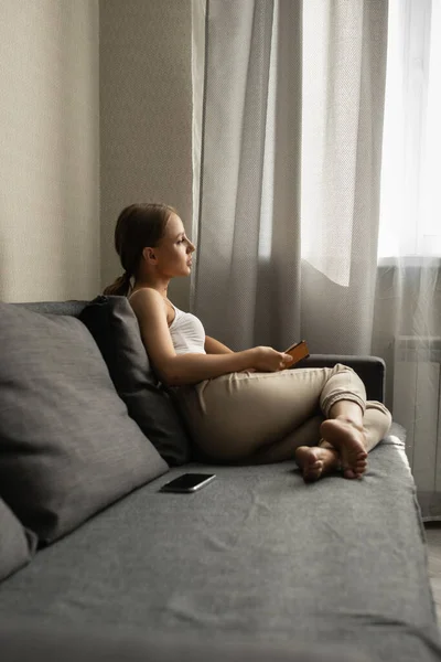 Une belle femme en pantalon et un haut est allongé sur le canapé, fini de lire un livre, regarde par la fenêtre — Photo