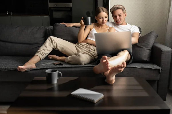 Krásná smějící se žena a muž sedí na pohovce, povídají si, pijí čaj a dívají se na film na notebooku — Stock fotografie