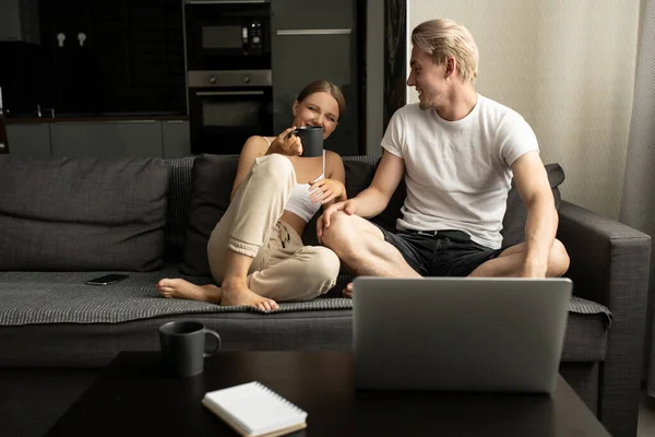 Žena a muž sedí na pohovce v bytě, povídají si, pijí čaj a dívají se na něco na notebooku. — Stock fotografie