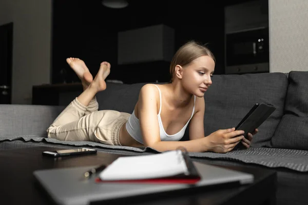 caucasian woman in casual clothes lies on the sofa at home, reads the news and checks notifications on the tablet