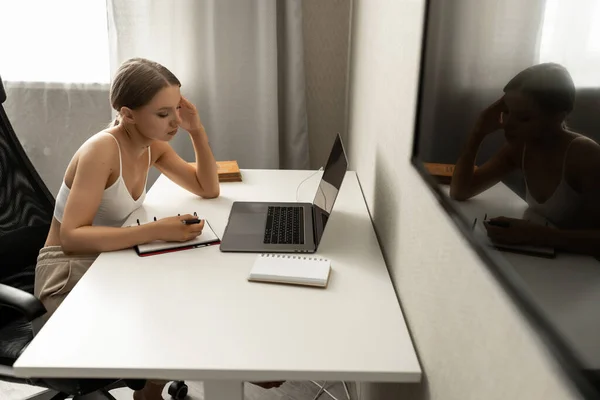 Mooie blanke vrouw in casual kleding zit op een stoel aan een werktafel thuis en maakt aantekeningen in een notebook. — Stockfoto
