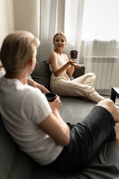 Krásná žena a muž sedí na pohovce, povídají si a pijí čaj. Příjemná konverzace mladého páru. — Stock fotografie