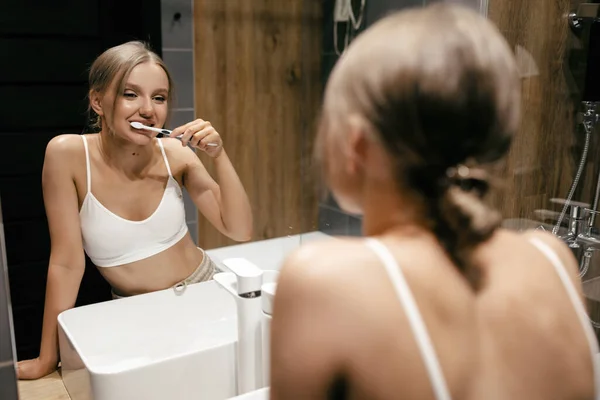 Mujer caucásica en el baño se cepilla los dientes con un cepillo sonriendo y mirándose en el espejo — Foto de Stock