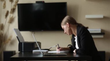 Kız evden uzaktan kumandalı bir bilgisayarla çalışıyor. Elinde not defteriyle. 