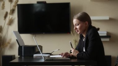 Kız evden uzaktan kumandalı bir bilgisayarla çalışıyor. Elinde not defteriyle. 