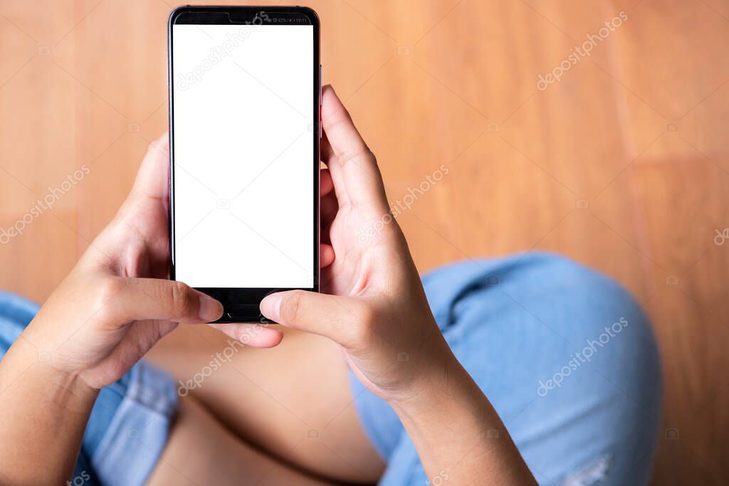 Top view mockup image of a woman holding  mobile phone with blank white screen while sitting on the wooden floor