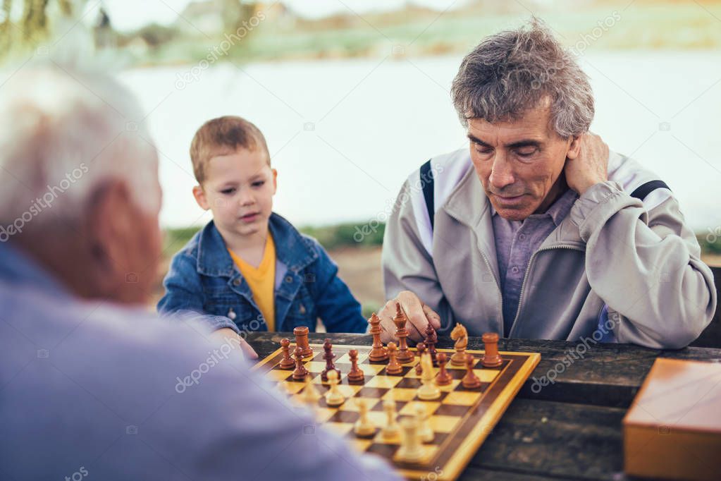 Active retired people, old friends and free time, two senior men having fun and playing chess at park, spend time with grandson