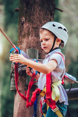 Küçük çocuk macera parkına kask ve güvenlik ekipmanıyla tırmanıyor.