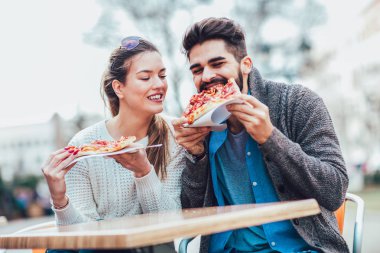 Dışarıda pizza yiyen ve gülümseyen bir çift. Açık bir kafede pizza yiyorlar..