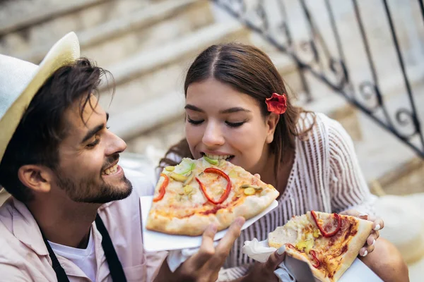 Happy couple of tourists eating tasty pizza on street sitting on stairs