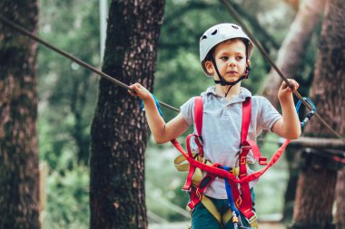 Küçük çocuk macera parkına kask ve güvenlik ekipmanıyla tırmanıyor.