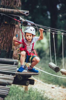 Küçük çocuk macera parkına kask ve güvenlik ekipmanıyla tırmanıyor.