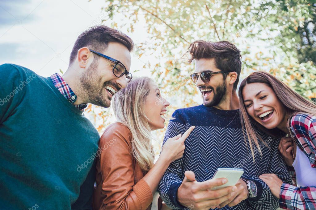 Image of four happy smiling young friends walking outdoors in the park holding smart phone