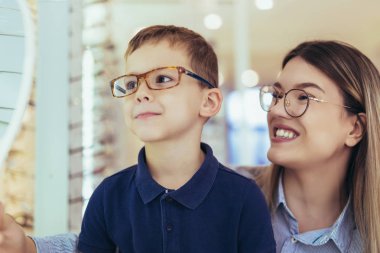 Mother with son choosing glasses in optics store. clipart