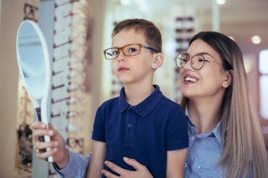 Mother with son choosing glasses in optics store. clipart