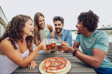 Grup Genç neşeli arkadaşıyla pizza zevk ve binanın çatısına masada otururken bira içmek