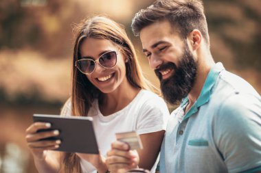 Bir parkın oturan genç çift tezgah ve online alışveriş için bir dijital tablet kullanır