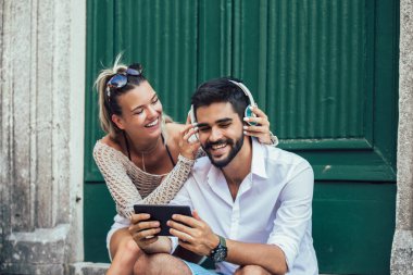 Tablet kullanma ve müzik merdivenlerde oturan güzel gülümseyen genç turist çift portresi.