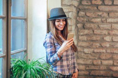 O pencereden dışarı bakarak akıllı telefon kullanan genç gülümseyen kadın portresi.