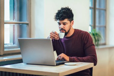 Bir fincan kahve tutarak ve dizüstü bilgisayar kullanarak rahat kıyafetler içinde yakışıklı Afro Amerikalı adam.