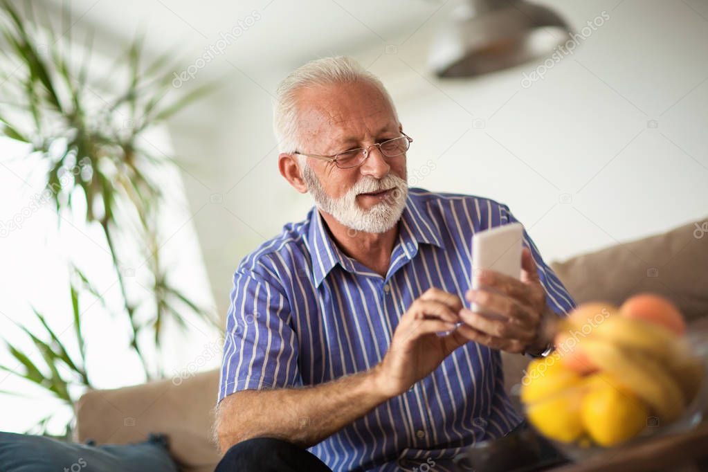 Handsome old man using a smartphone and smiling while sitting on couch at home