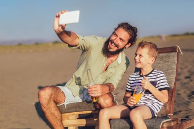 Baba ve oğul selfie plajda alarak bir kadeh. Genç adamla küçük çocuk kendi kendine portre deniz sahilinde akıllı telefon kullanarak yapma.