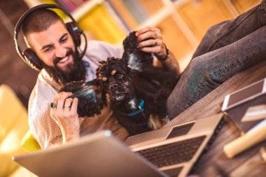 Evde dizüstü bilgisayarda çalışan yakışıklı ve dövmeli bir adam masada sevimli bir köpekle oturuyor.