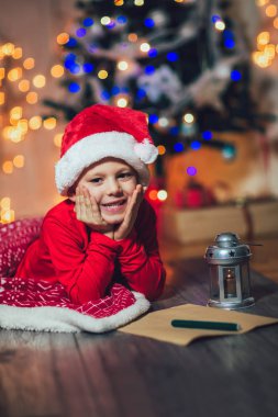 Çocuk Noel ağacının yanındaki kırmızı şapkalı Noel Baba 'ya mektup yazıyor.