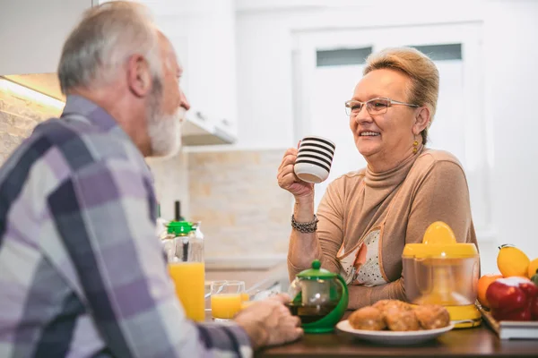 Elderly couple in the kitchen preparing breakfast. They drink coffee or tea and talk