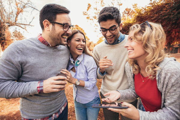 Four happy smiling young friends walking outdoors in the park holding digital tablet