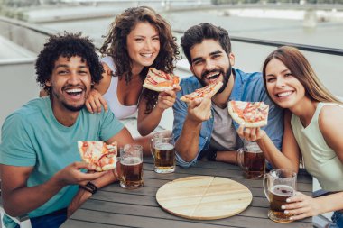 Arkadaşlar pizzayı seviyor. Bir grup neşeli insan pizza yiyip bira içerken binanın çatısındaki fasulye çuvallarının başında oturuyorlar.