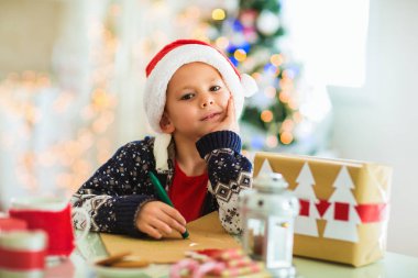 Çocuk Noel ağacının yanındaki kırmızı şapkalı Noel Baba 'ya mektup yazıyor.