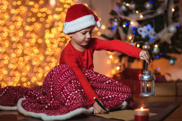 Boy writing letter to Santa Claus in red hat near the Christmas tree