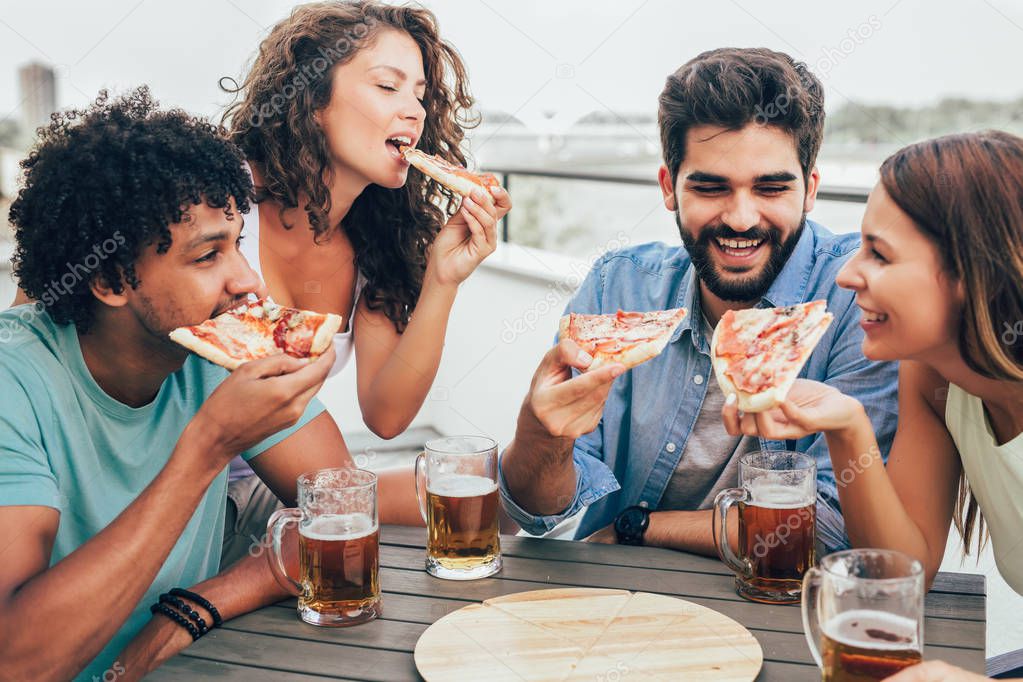 Friends enjoying pizza. Group of young cheerful people eating pizza and drinking beer while sitting at the bean bags on the roof of the building