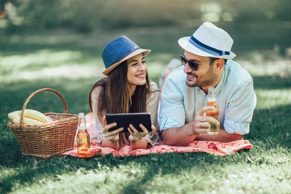 Couple lying on a picnic blanket in a park with a picnic basket filled with fruit, they are using digital tablet