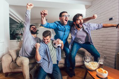 Erkekler televizyonda birlikte spor izleyip neşeli çığlıklar atıyorlar. Bir grup arkadaş kanepede oturup futbol maçı izliyorlar..