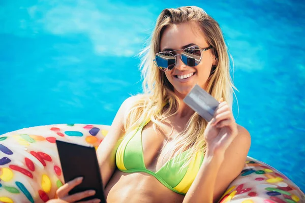 使用数字平板电脑和信用卡 在游泳池的充气床垫上享受晒黑的女人 — 图库照片