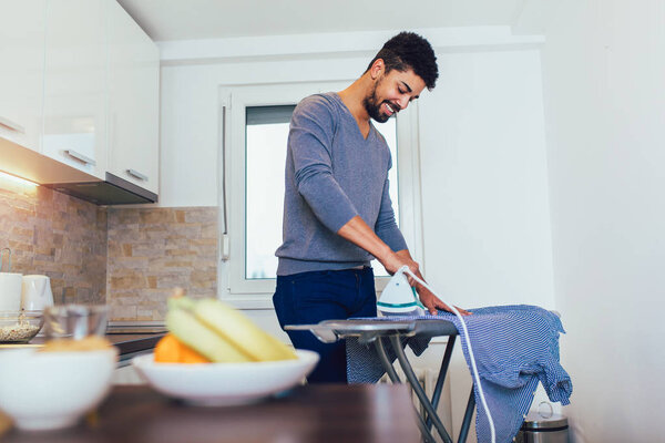 Attractive american black man is ironing shirt at home. 