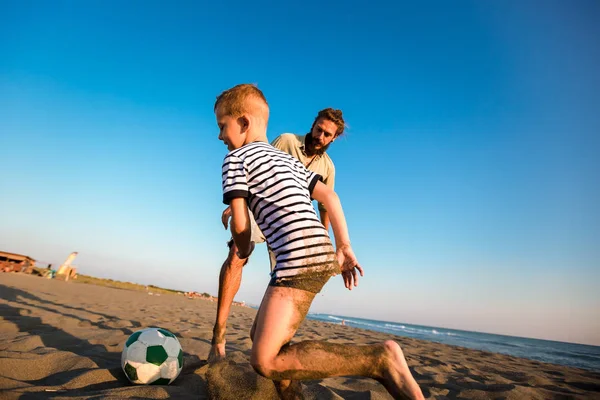Счастливые отец и сын играют в футбол или футбол на пляже, имея — стоковое фото