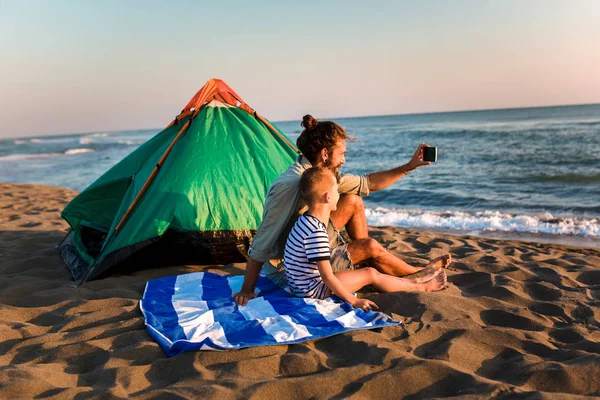 Far og sønn på stranden bruker telefon og har det gøy. – stockfoto