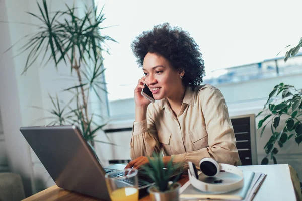 Leende unga afrikanska kvinnliga entreprenör sitter vid ett skrivbord i h — Stockfoto