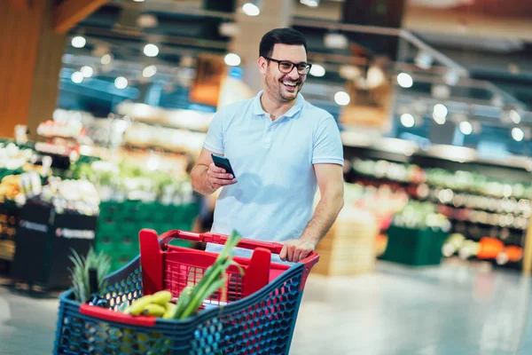 Handsome man shopping in supermarket pushing trolley and holding — Stock Photo, Image