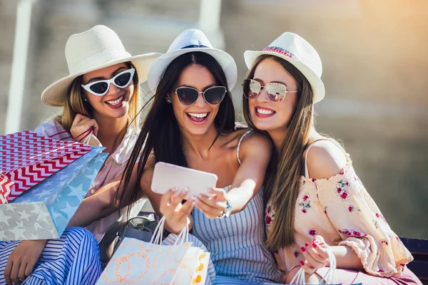 Tre ragazze con borse colorate per la spesa utilizzando smartphone — Foto Stock