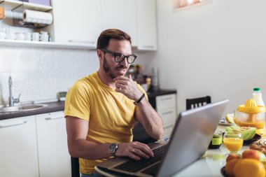 Evde çalışan meşgul adam, kahvaltı yaparken laptop kullanarak.