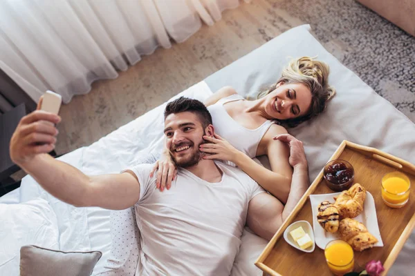 Joven pareja feliz desayunando en habitación de hotel de lujo usando s — Foto de Stock