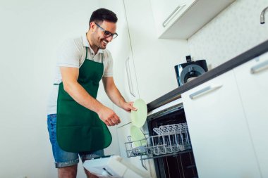 Adam mutfakta ev işi yapıyor, bulaşıkları bulaşıkmakinesine dolduruyor.