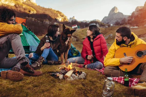 Grupo de amigos sorridentes sentados em torno da fogueira no acampamento — Fotografia de Stock